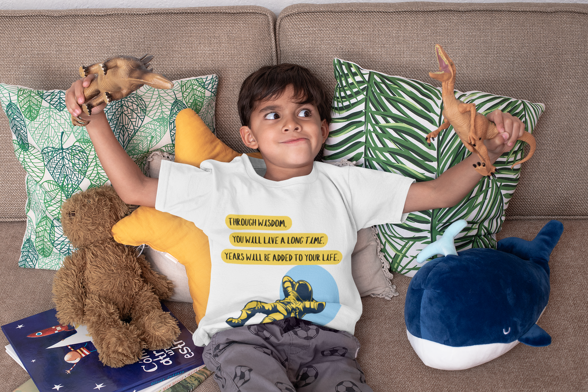 Child sitting on a couch with stuffed animals and a book, wearing a white t-shirt with text and graphics.