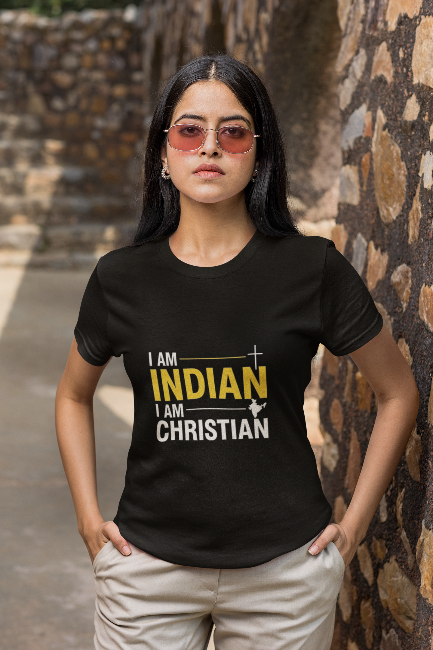 Person wearing a black t-shirt with 'I AM INDIAN I AM CHRISTIAN' text, standing against a stone wall.