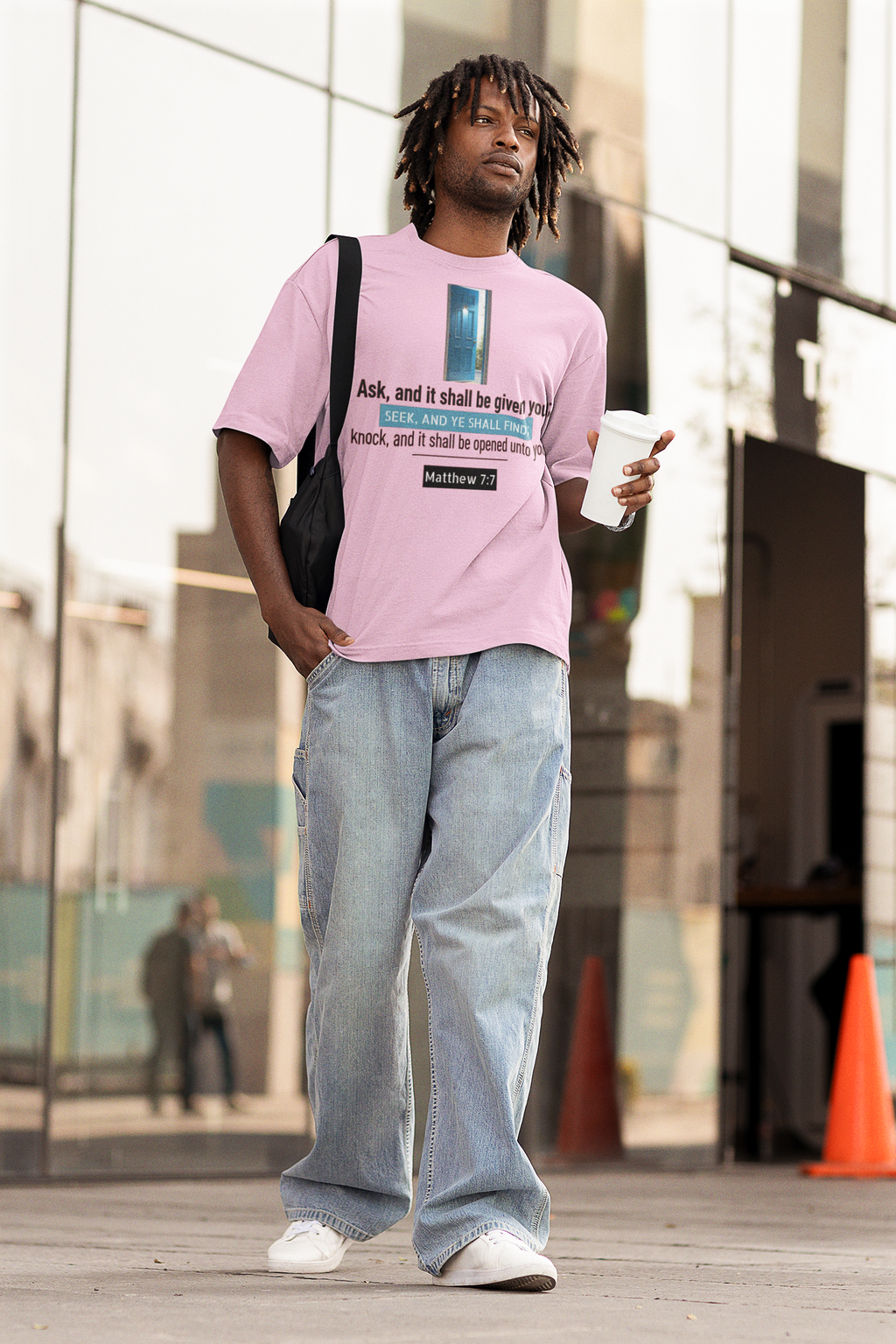 Man wearing a pink t-shirt with text and light blue jeans, holding a coffee cup, standing in an urban setting.
