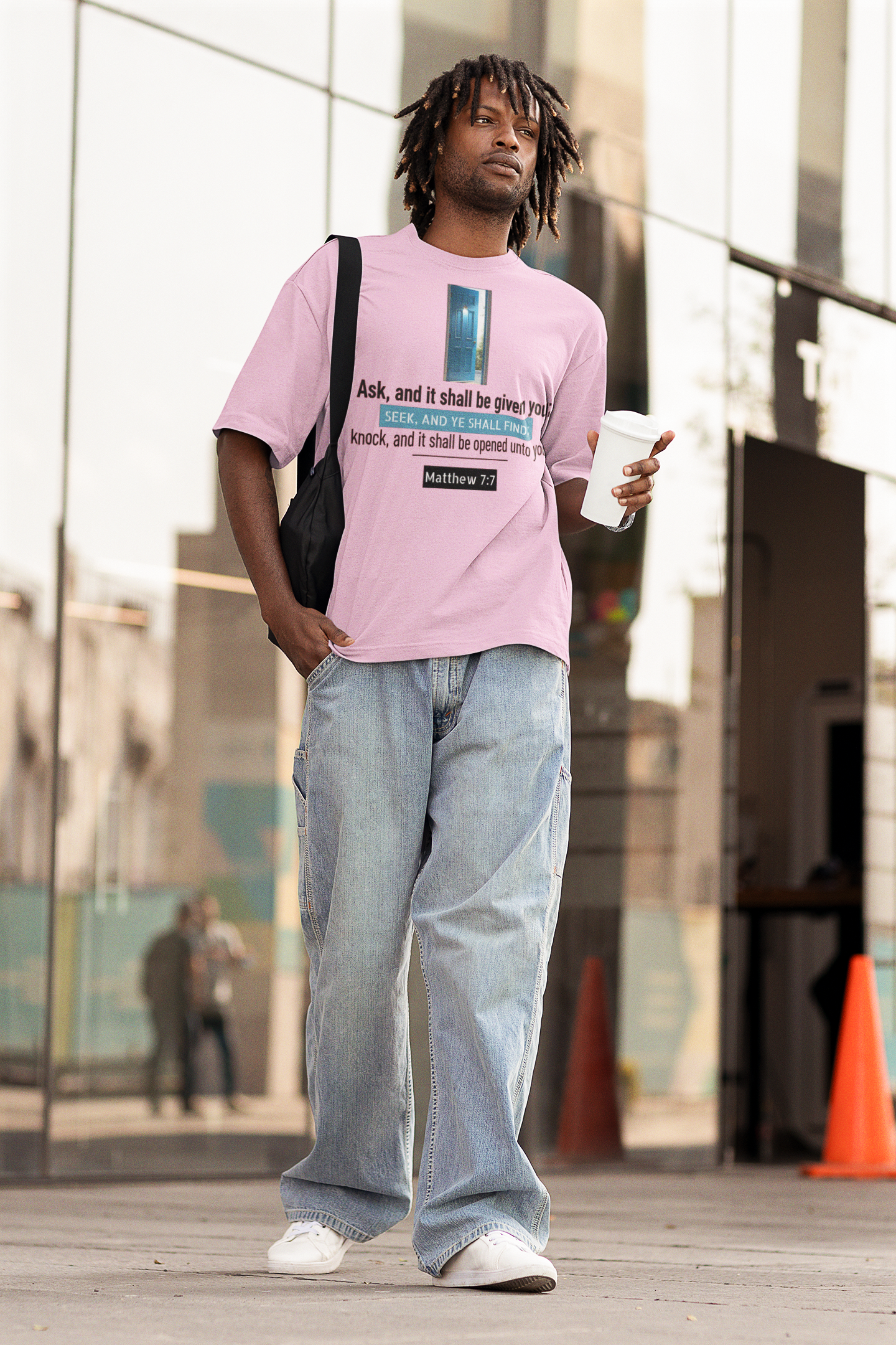 Man wearing a pink t-shirt with text and light blue jeans, holding a coffee cup, standing in an urban setting.