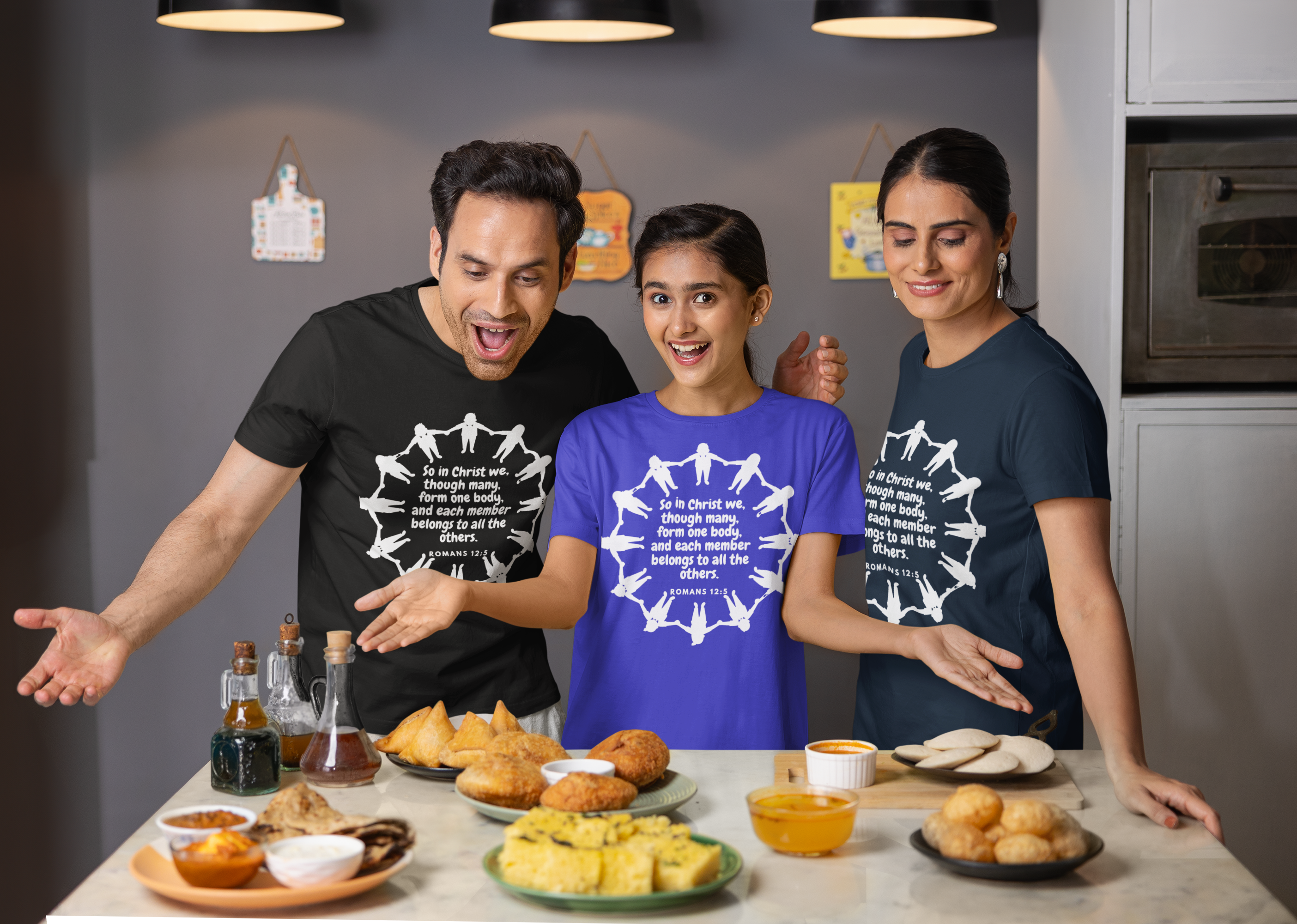 Three people wearing t-shirts with a design, standing around a table with food items.