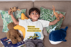 Child sitting on a couch with stuffed animals and a book, wearing a white t-shirt with text and graphics.