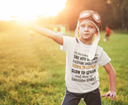 Child wearing a white t-shirt with text and goggles, standing in a grassy field with a warm glow.