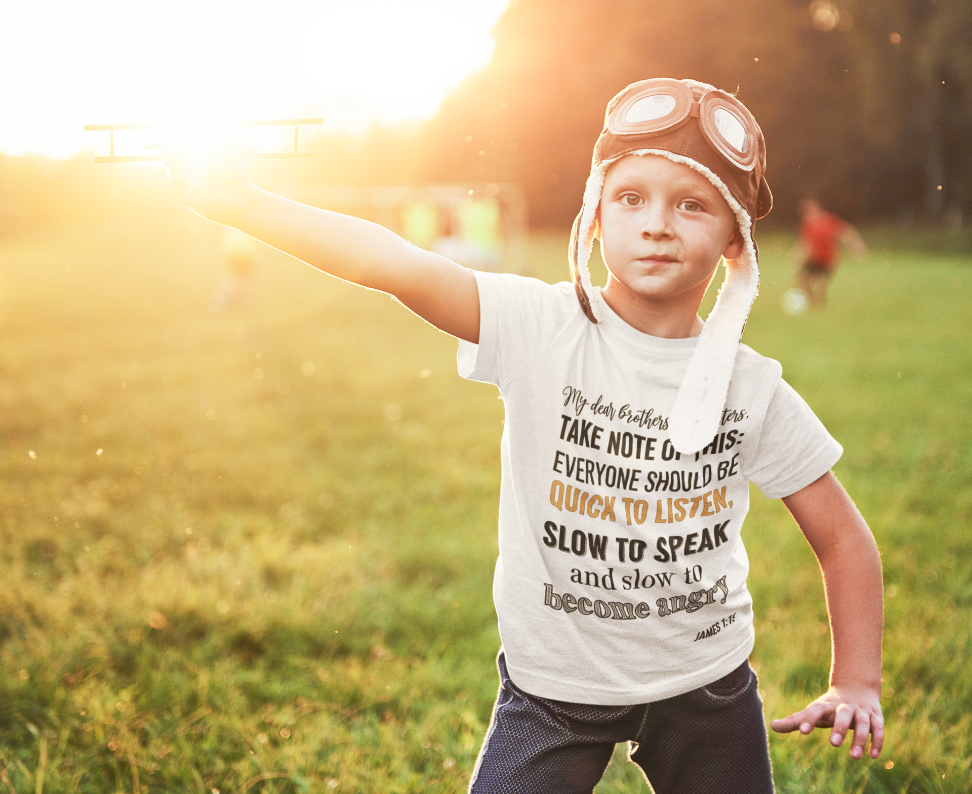 Child wearing a white t-shirt with text and goggles, standing in a grassy field with a warm glow.
