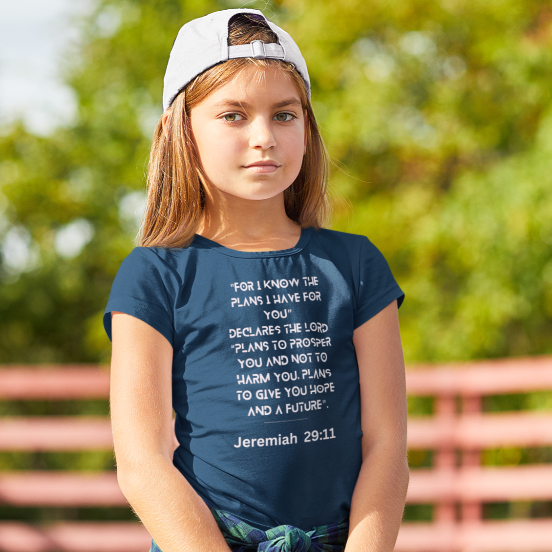Young girl holding a pink skateboard with a blue t-shirt and white cap outdoors.