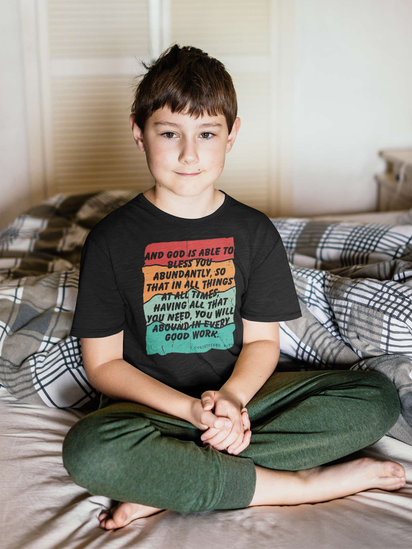 Young boy sitting on a bed wearing a black t-shirt with colorful text.