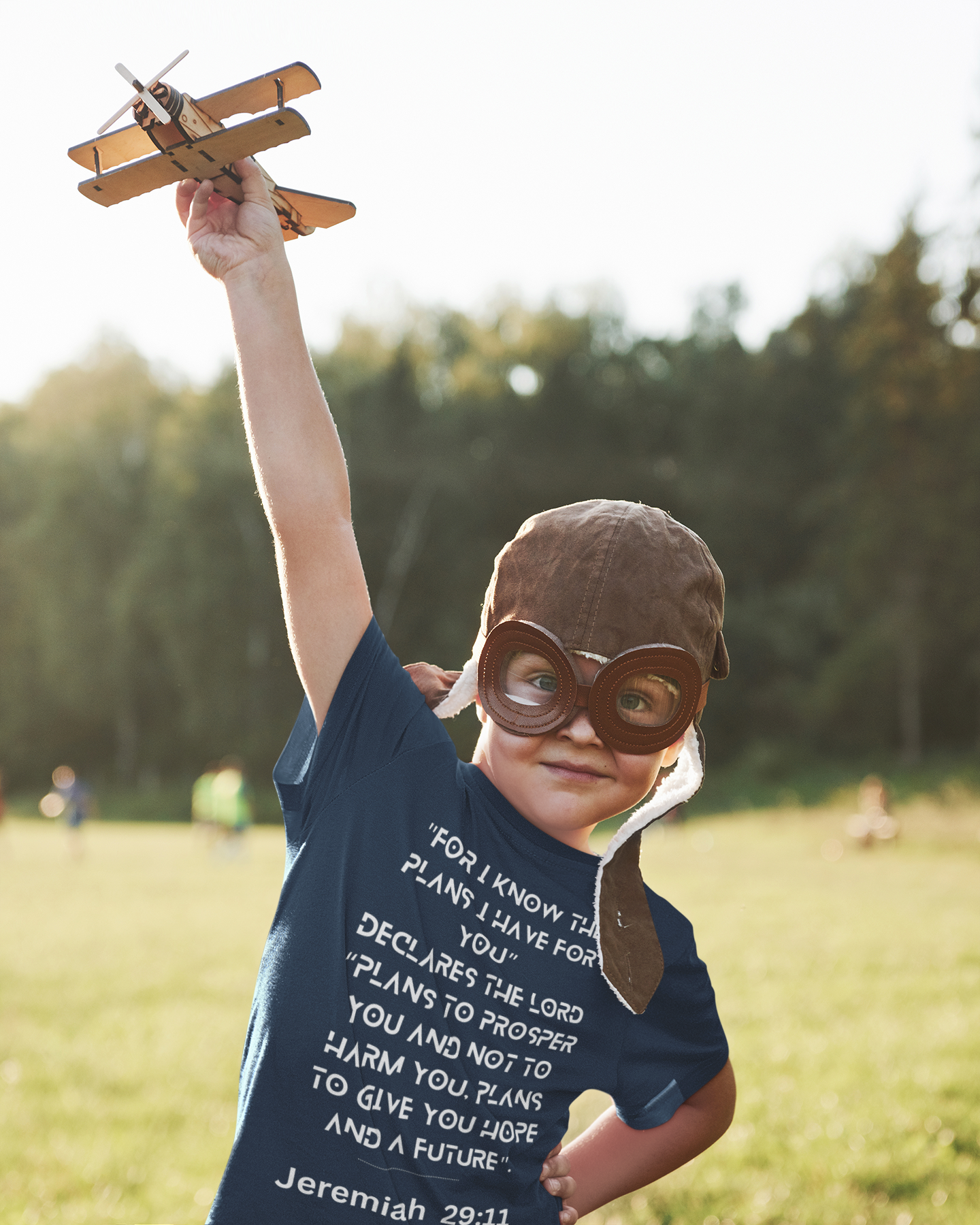 Child in a field wearing an aviator hat and holding a model airplane, with a scenic background.